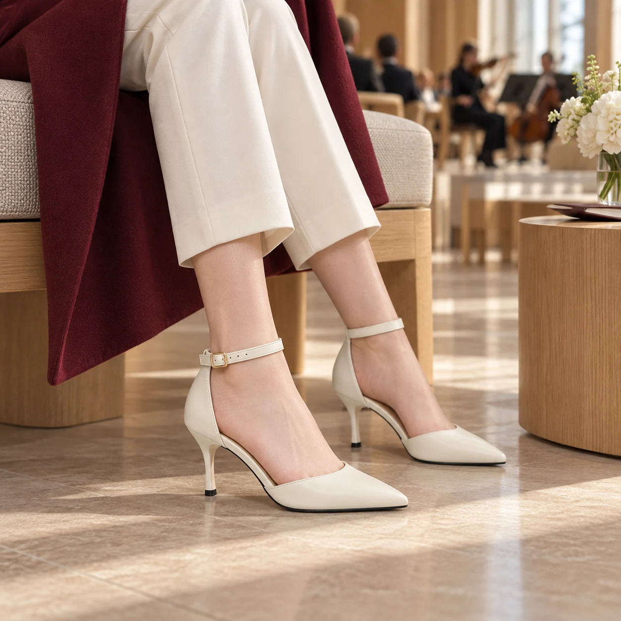 Petite woman seated in Ivory Grace heels with ivory trousers and a burgundy coat at a concert hall